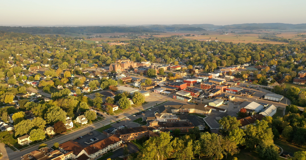Aerial view of a small rural American town surrounded by farmland and rolling hills, with tree-lined streets, a downtown commercial district, and residential neighborhoods visible at golden hour.